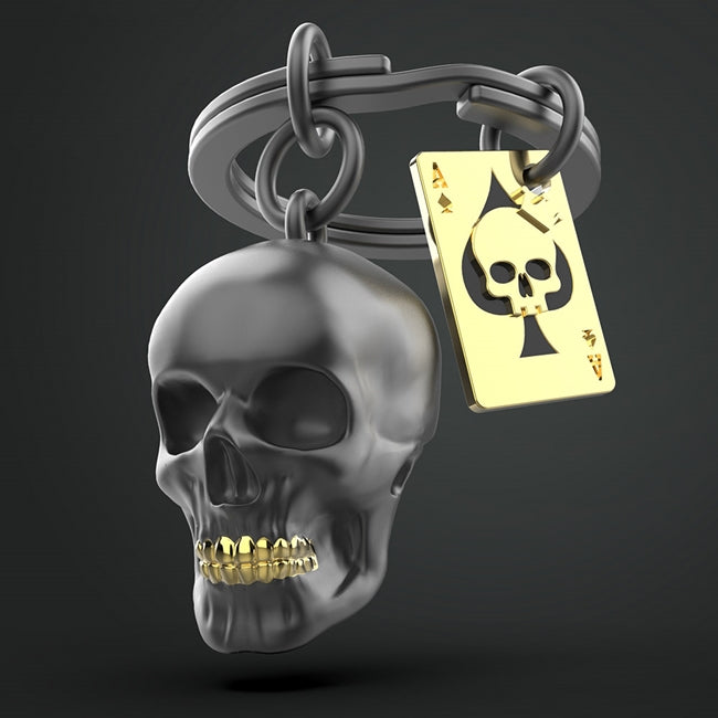 Metalmorphose.lt metalinis raktų pakabukas KAUKOLĖ IR KORTA. Tvirtas metalinis korpusas, juodai nudažyta kaukolė, auksinė korta, detalizuota kaukolės 3D miniatiūra. Nerūdijančio plieno žiedas. Pakabukas iš KASDIENYBĖS PASAULIS kolekcijos.