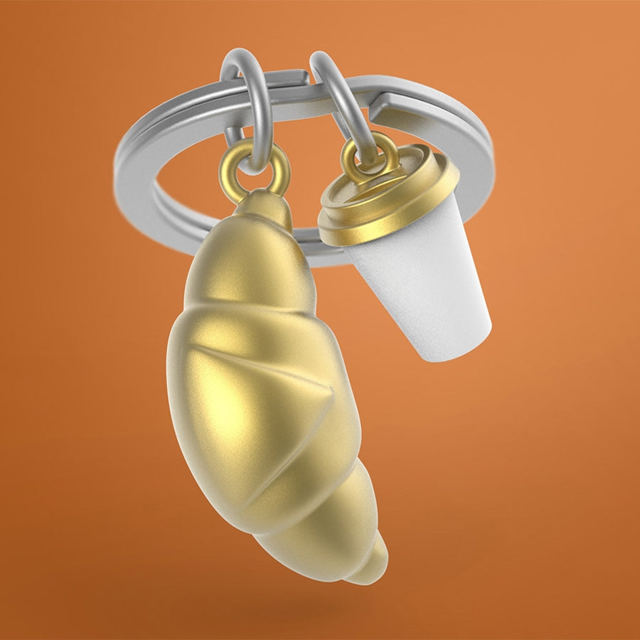 Metalmorphose metalinis raktų pakabukas „Pasimatymas Paryžiuje“ – 3D kruasano ir kavos puodelio dizainas iš Skonių Pasaulis kolekcijos. Detalės auksinis dangtelis, auksinė kruasano tekstūra, plieninis žiedas