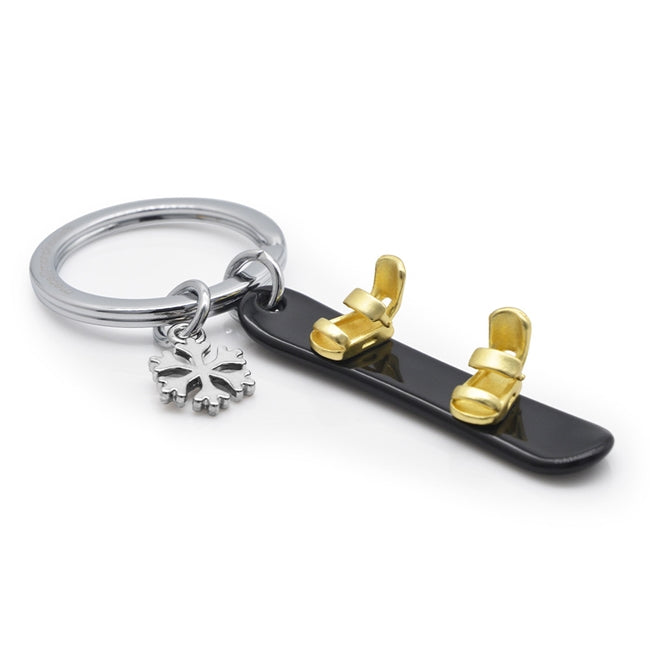 Metalmorphose.lt metalinis raktų pakabukas SNIEGLENTĖ IR SNAIGĖ. Juoda 3D snieglentė su auksiniais segtuvais, šalia spindinti metalinė snaigė, premium cinko lydinys, plieno žiedas. SPORTO PASAULIS kolekcija.